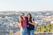 Two smiling women pose on a scenic overlook with a city view behind them.