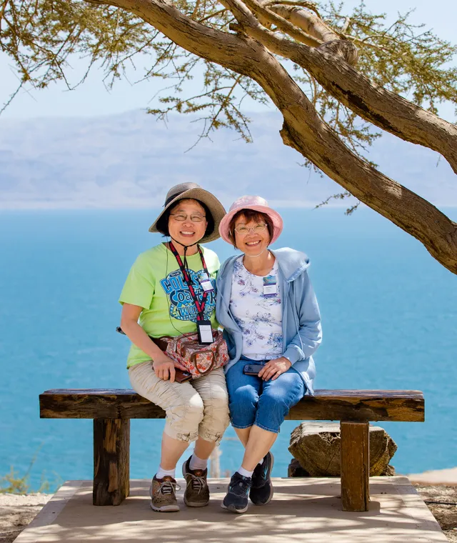 Two smiling women sitting on a wooden bench with a scenic ocean view behind them