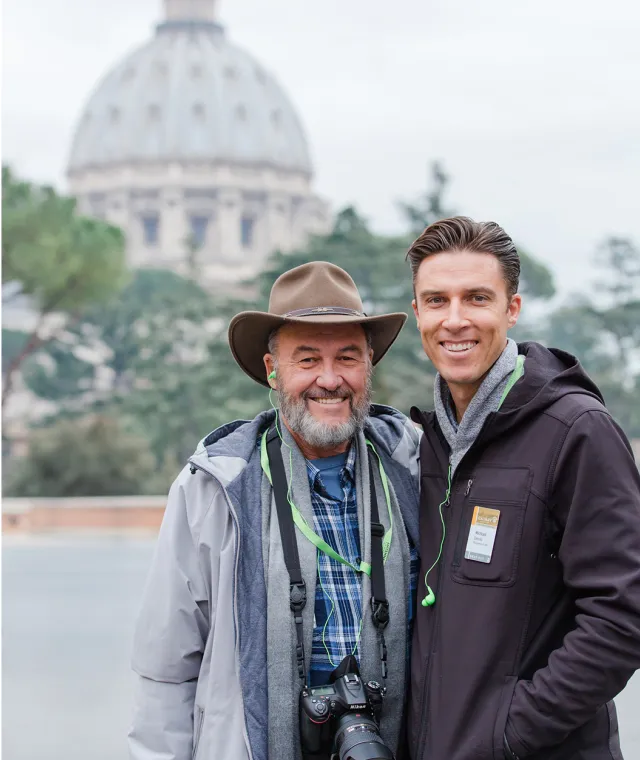 Two men standing together with trees and a domed architectural structure behind them
